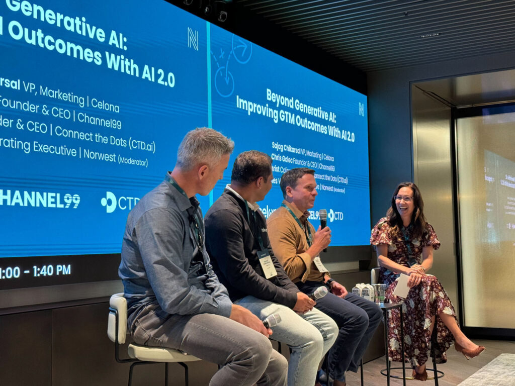 A panel of four speakers sitting on a stage in front of two large blue screens