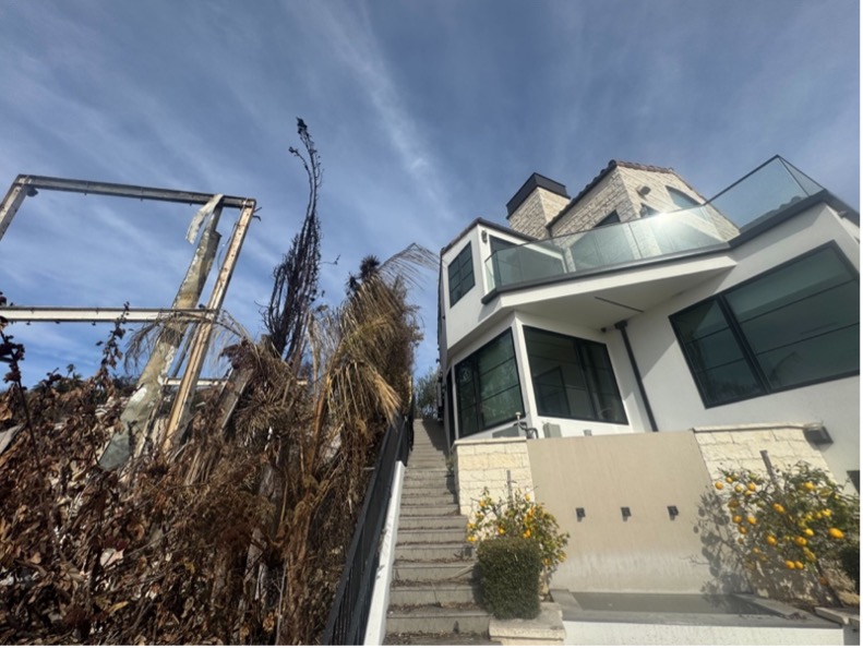 Photograph of a modern hillside home with intact windows and clean white walls, contrasted with scorched trees and burned remains of a neighboring structure. Caption context: the home survived a wildfire thanks to the Frontline Wildfire Defense system, standing as the last structure on the street.