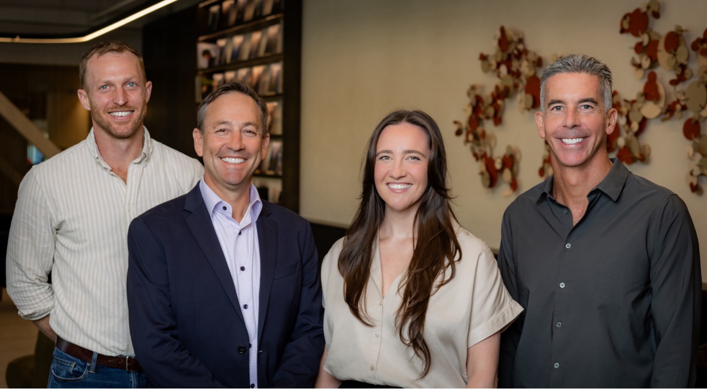 Group photo of four people standing indoors, smiling at the camera. From left to right: a man in a light striped shirt, a man in a navy blazer and white shirt, a woman in a beige blouse, and a man in a dark gray button-up shirt. Caption context: Norwest Partner Scott Mitchell, Frontline CEO and Founder Harry Statter, Norwest Vice President Cassie McHenry, and Norwest Managing Partner Jon Kossow.