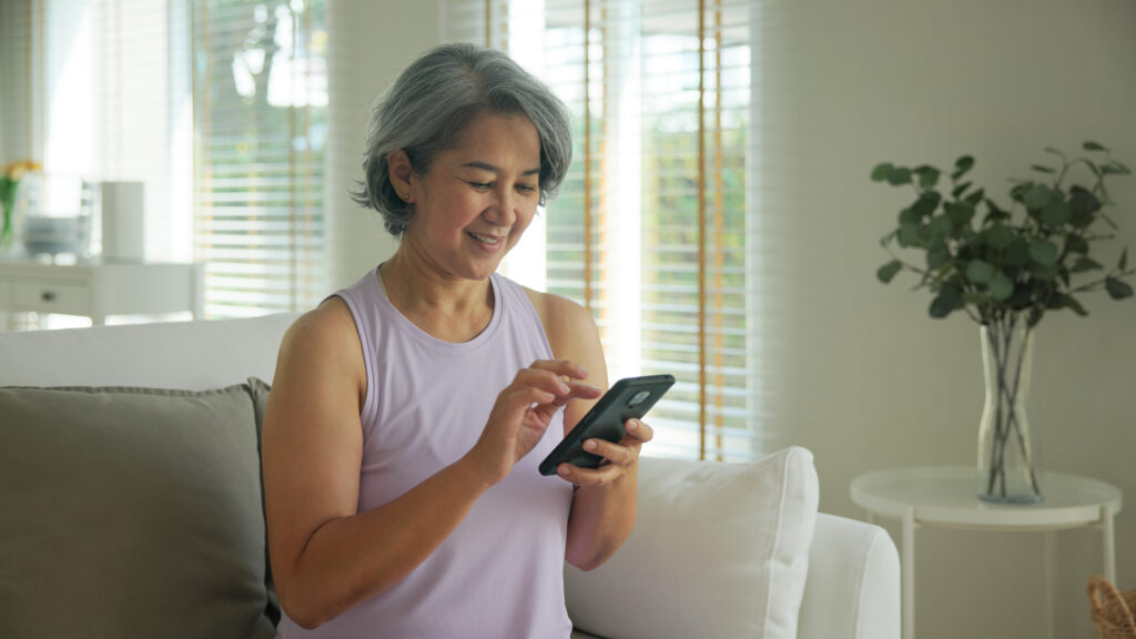 Smiling woman with gray hair uses her smartphone while sitting on a sofa in a bright living room.