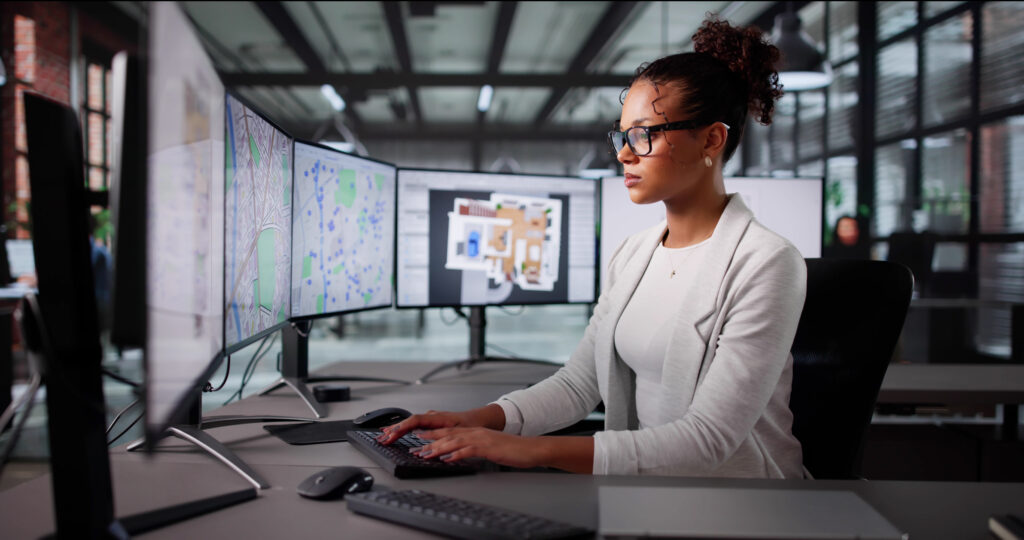 Woman analyzing maps and floor plans on multiple monitors in a modern office.